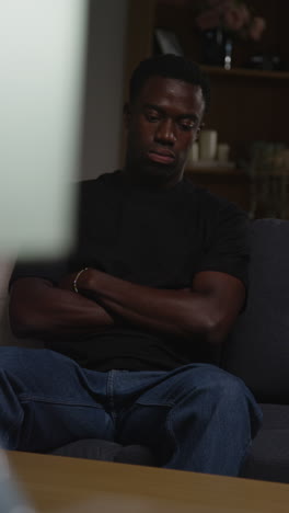 vertical video of unhappy and depressed young man sitting on sofa at home looking anxious and worried resting head on hand