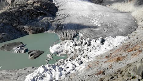 Aerial-flyover-over-the-lake-of-Rhone-glacier-near-Furka-mountain-pass-at-the-border-of-Valais-and-Uri-in-Switzerland-with-a-pan-up-towards-the-ice-mass-and-mountain-peaks-on-a-summer-day