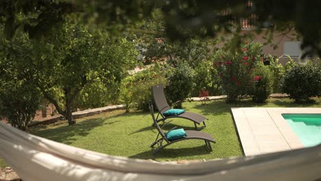 dos sillas de descanso con cojines azules frente a un jardín exuberante y una piscina, vistas a través de una hamaca