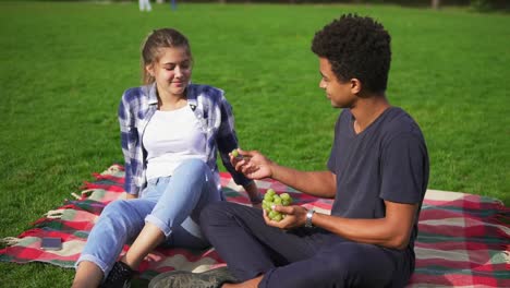 Junges-Schönes-Paar,-Das-An-Einem-Sommertag-Auf-Dem-Gras-Im-Park-Obst-Isst.-Mann-Füttert-Frau-Sorgfältig-Mit-Saftigen-Trauben