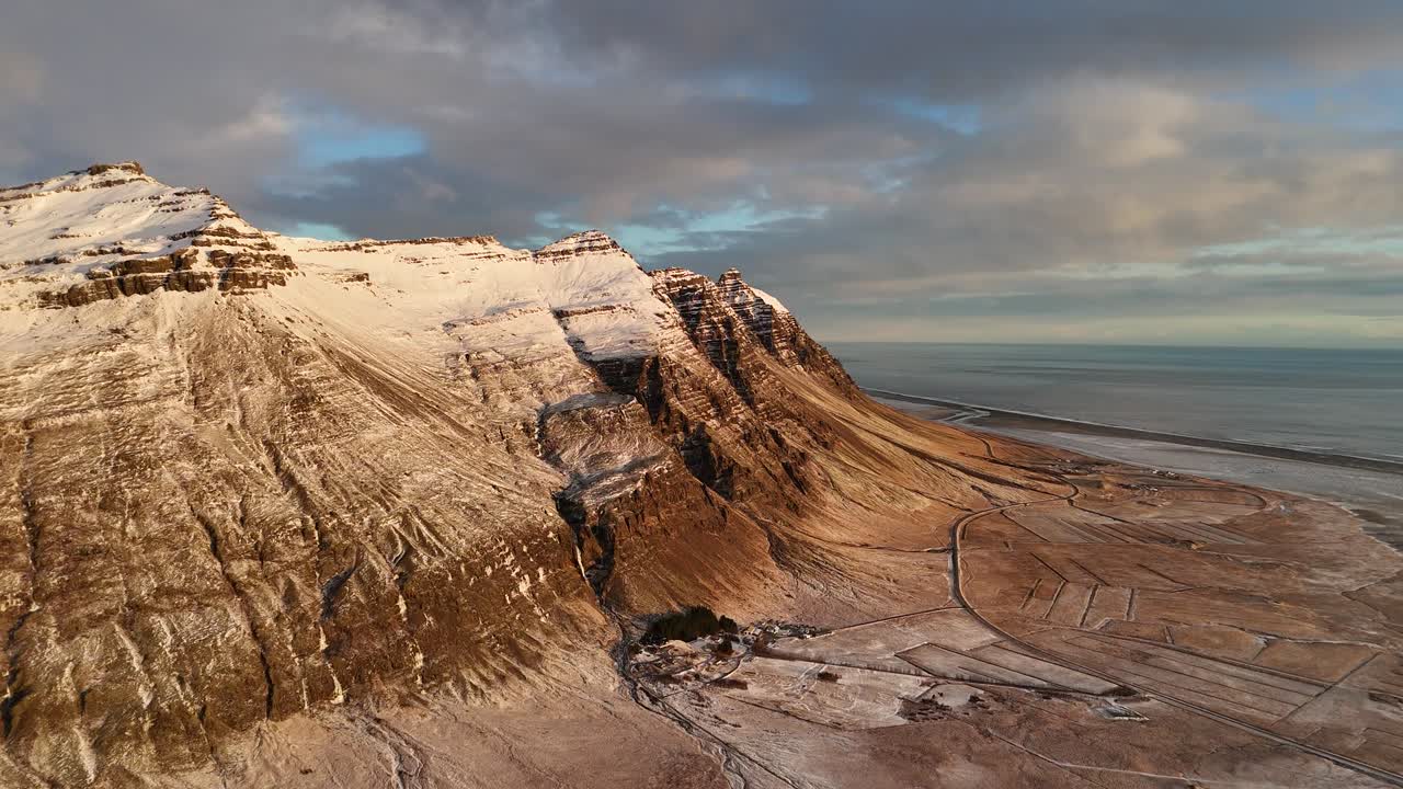 Aerial drone view of dramatic snow-capped mountains beside Icelandic coastline during winter. Rugged peaks, icy slopes, and golden light create contrast over remote landscape near frozen shoreline