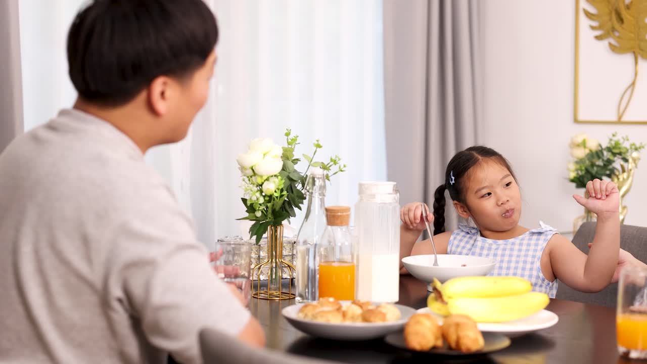 Young girl eats breakfast with parents, natural morning light, cheerful mood, static camera angle