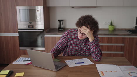 curly haired male student attractive young boy in glasses is studying at home using laptop typing writing in notebook. college student using laptop computer watching distance online learning seminar