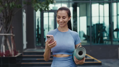 woman in sportswear using a phone while carrying a yoga mat outside a gym