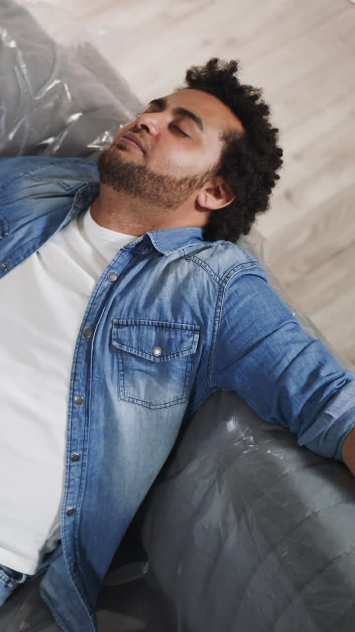 Exhausted black man falls down and takes off glasses on foiled sofa upper view. Finishing repair process in house. African-American man rests after work