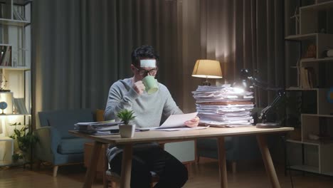 아픈 아시아인 남자가 집에서 문서와 함께 열심히 일하면서 물을 마시고 있습니다.