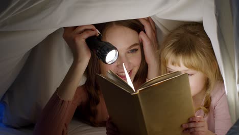 young mother reading goodnight story fairytale to child daughter under duvet blanket in night room