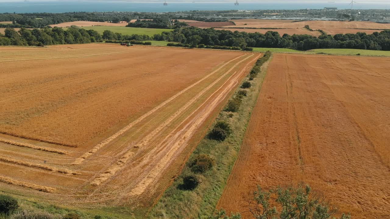 Drone shot of harvester in field with with sea, wind turbine and rigs in the distance