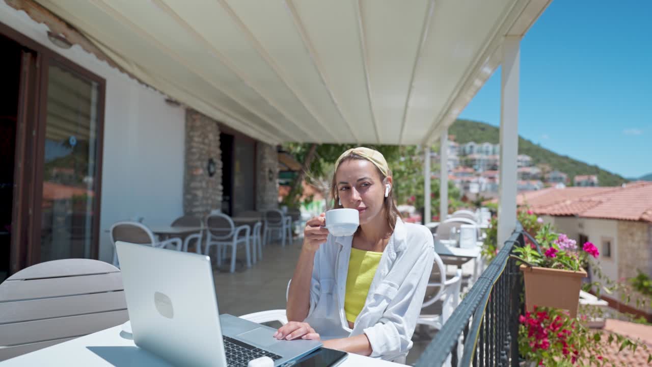 Pleased woman enjoying morning coffee while sitting on terrace with sea view on Turkish resort, using laptop computer for remote work, freelance concept
