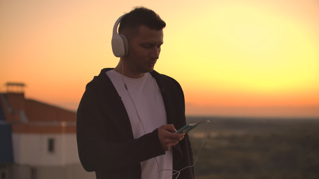 A man in big white headphones listens to music standing on the roof at sunset. Relaxation.