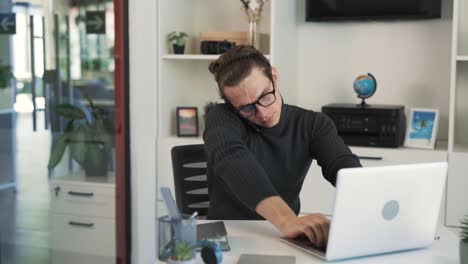 A-very-busy-man-sits-at-his-work-desk,-talking-on-the-phone,-typing-on-his-laptop,-and-taking-notes-in-his-notebook