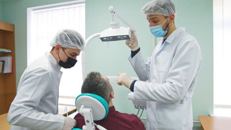 two male students of dentistry department practicing on a patient. one student holding and directing a lamp over the man in chair.