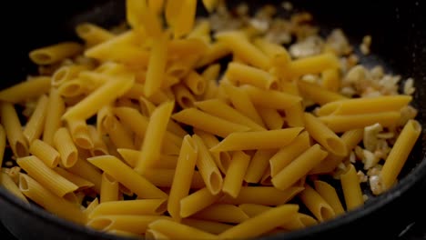 cocinero anónimo poniendo macarrones crudos en una sartén con cebolla frita