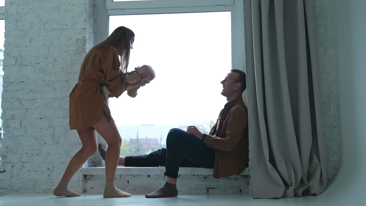Happy smiling Caucasian man sitting on the window-sill. Woman standing beside waving a baby and then sits down near her husband.
