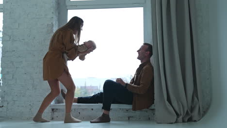 happy smiling caucasian man sitting on the window-sill. woman standing beside waving a baby and then sits down near her husband.