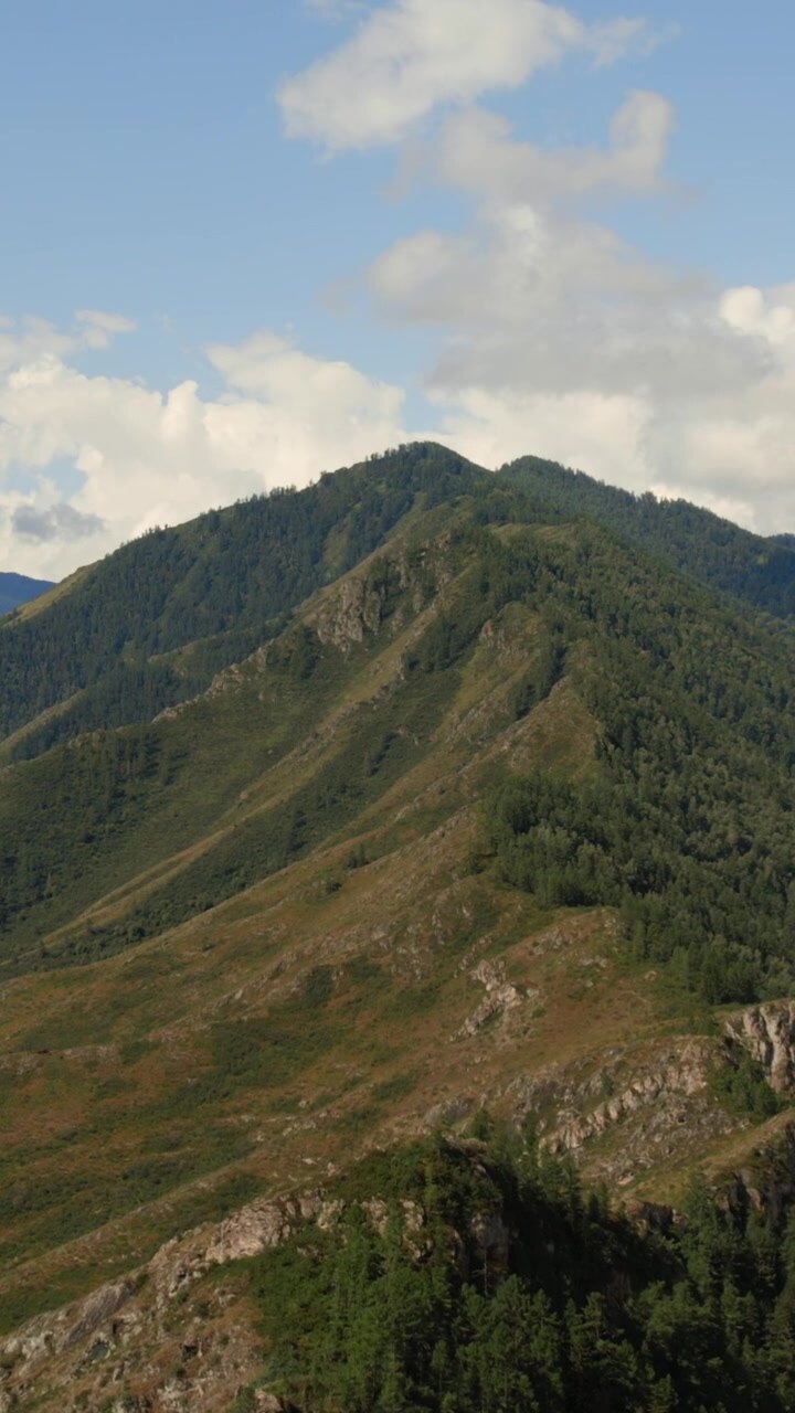 Toma amplia de una cresta montañosa verde y laderas rocosas bajo un cielo azul con nubes en el Valle de Ak-Kem, República de Altái, Rusia, una serena escena de naturaleza salvaje para viajes.