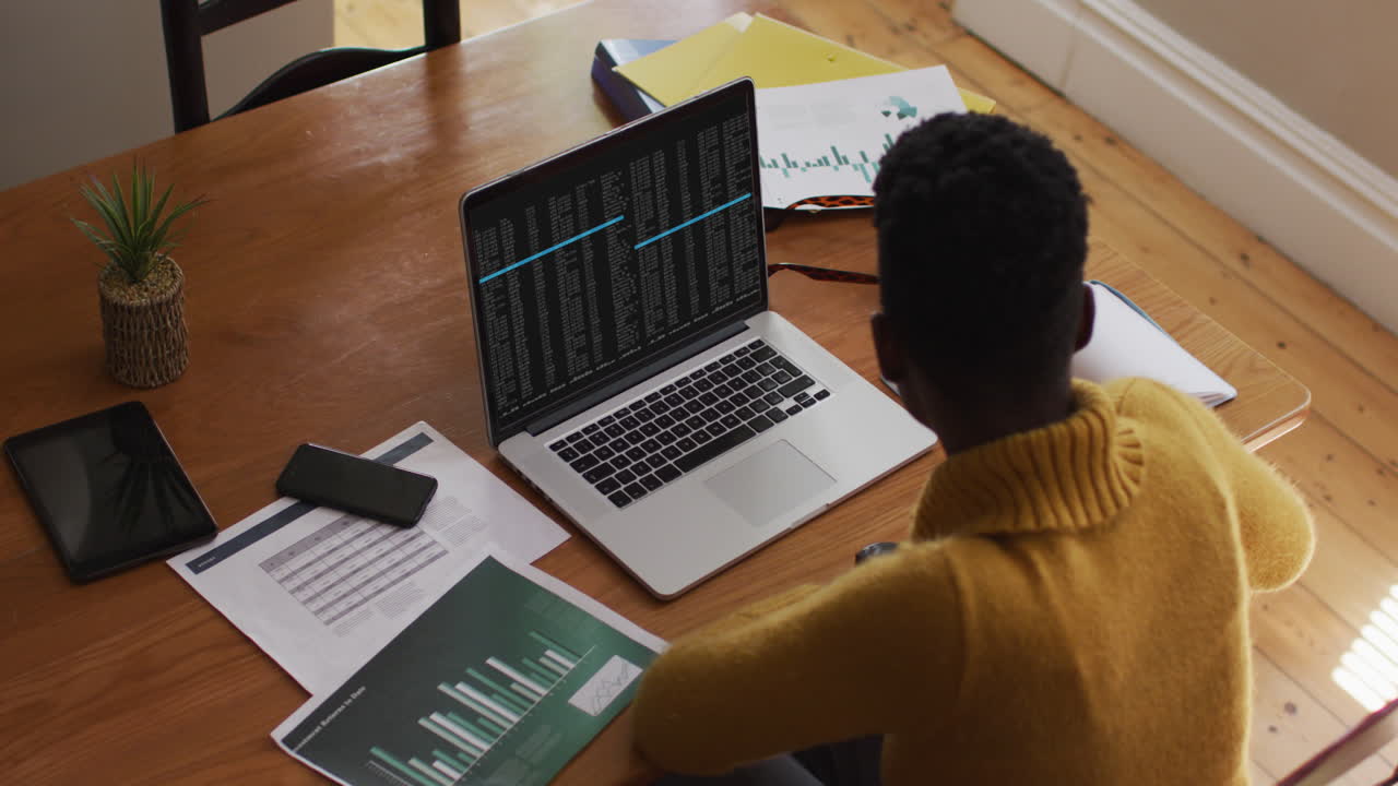 African american man sitting at desk watching coding data processing on laptop screen