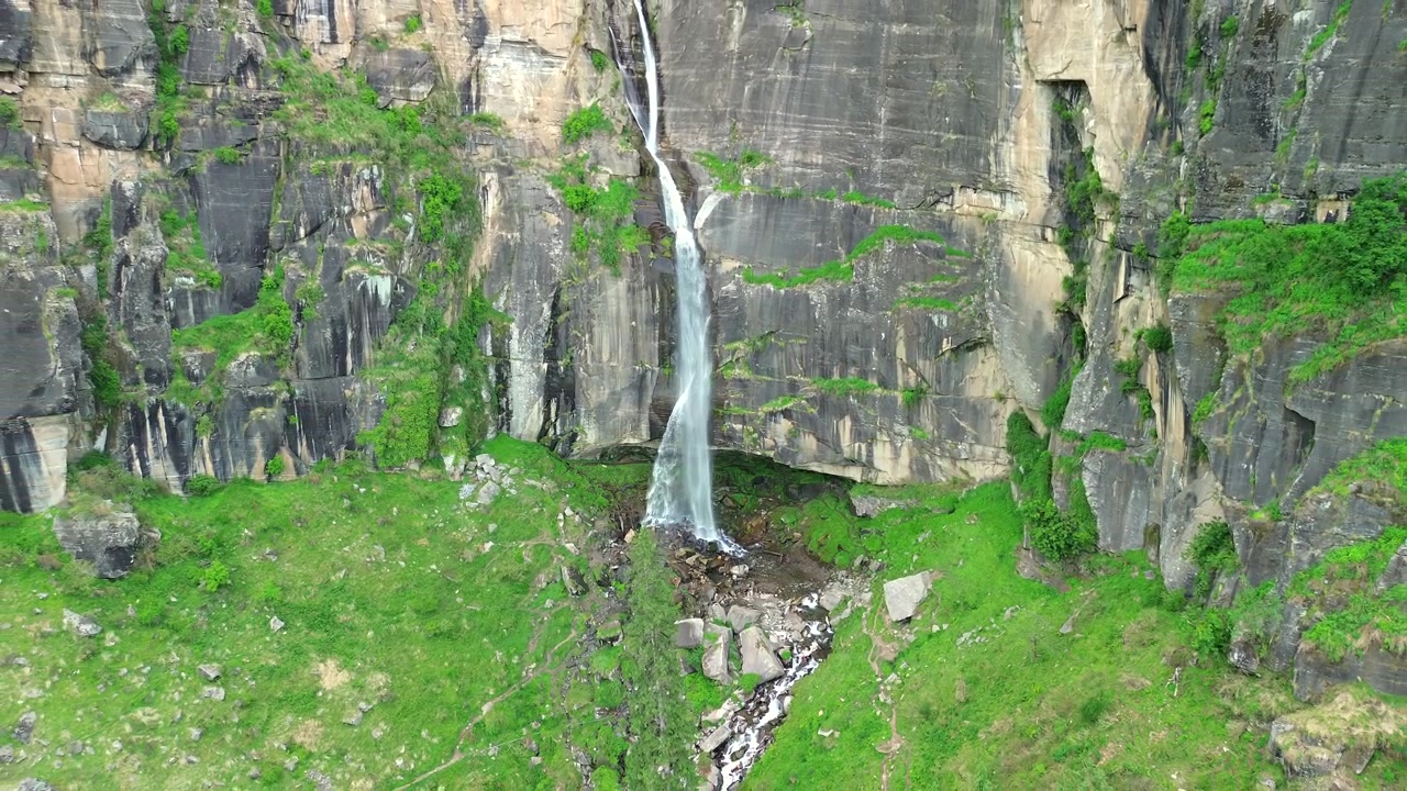 Ripresa lenta con drone che si allontana, rivelando una maestosa cascata stretta che si getta da un'alta scogliera verticale di pietra nera, circondata da chiazze di erba verde a Manali, Himachal Pradesh, India.