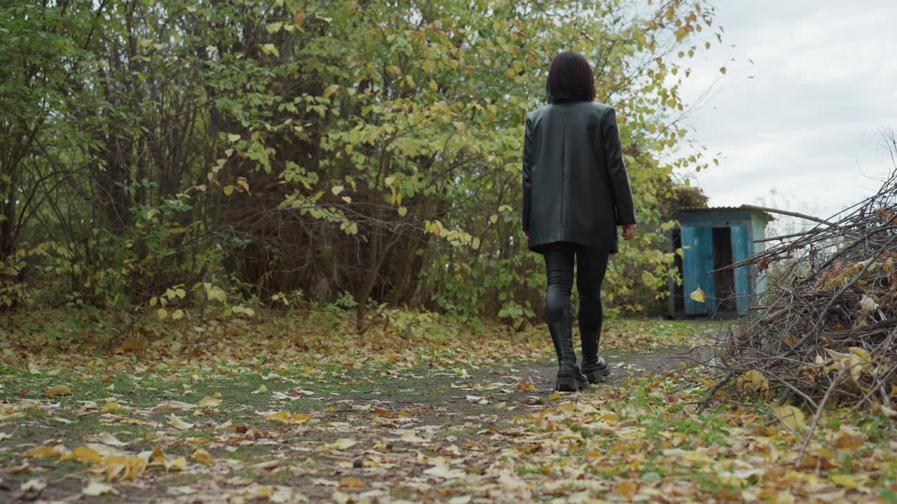 Donna con giacca di pelle che cammina lungo il sentiero di un bosco autunnale verso una rimessa blu, foglie cadute che scricchiolano sotto gli stivali, cielo nuvoloso che proietta una luce malinconica, atmosfera da rocker solitario, passo sicuro, texture della pelle