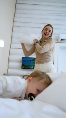 toddler boy sucking a pacifier lies on the bed. blonde woman taps a baby with a pillow playfully. mother and happy son game at home. vertical video.
