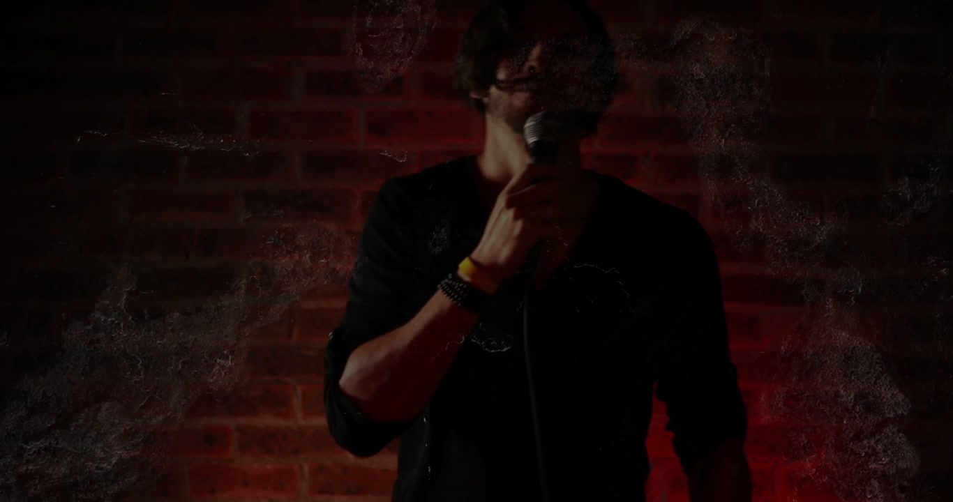 Male performer raising wired mic, starting music on red brick wall, film grain pulsing around him