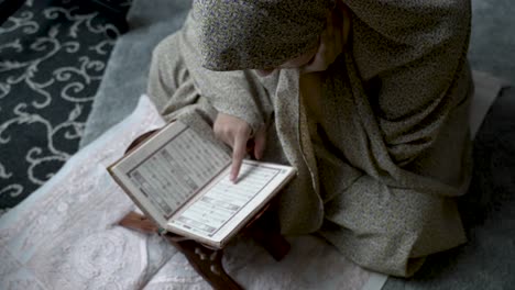 seated on the ground amidst stacked clothing, a young muslim girl immerses herself in a religious text, embodying the notion of studying and acquiring knowledge in the cozy confines of home