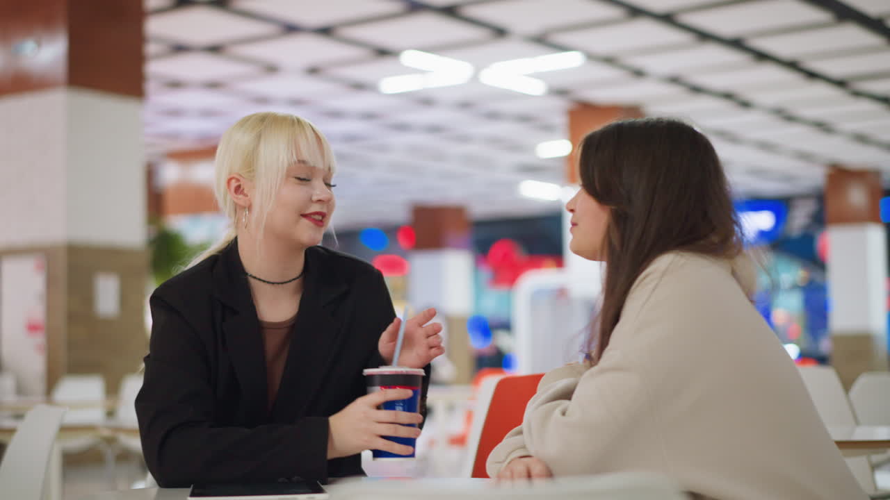 Girl extends drink to friend sitting at table in modern mall cafe with bright interior, casual friendly gesture showing moment of sharing and connection in relaxed social setting