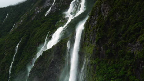 Tilt-up-revealing-high-waterfalls-of-Milford-Sound-in-moody-cloudy-weather,-New-Zealand