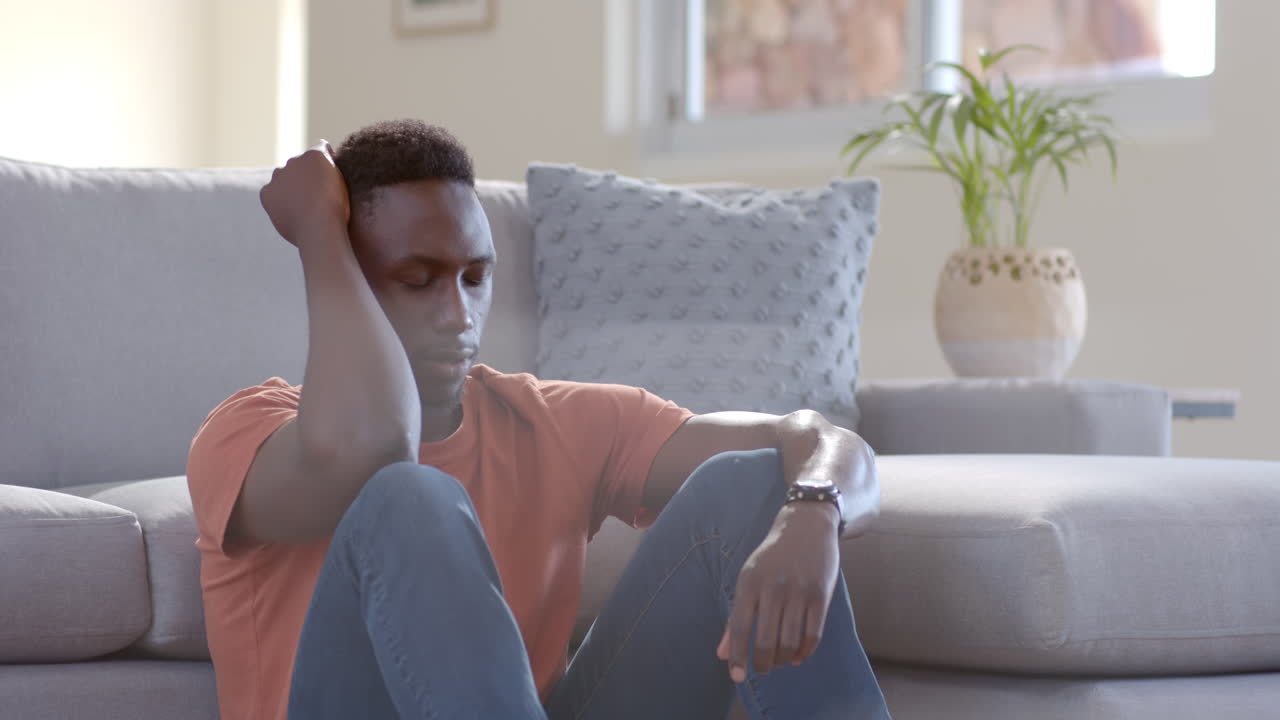 Worried african american man sitting and thinking in sunny living room, slow motion