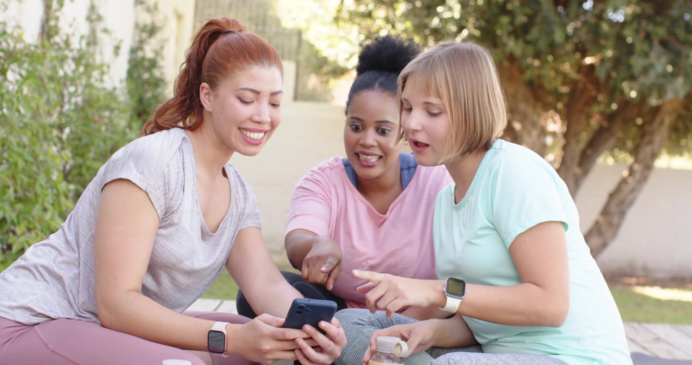Diverse women on yoga mats in yard, left woman showing phone, others reacting and sharing laughs