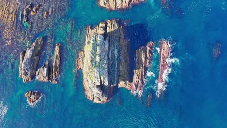 Aerial-vertical-high-angle-view-of-big-rocks-in-sea-coast-while-waves-break-against-them-at-sunset