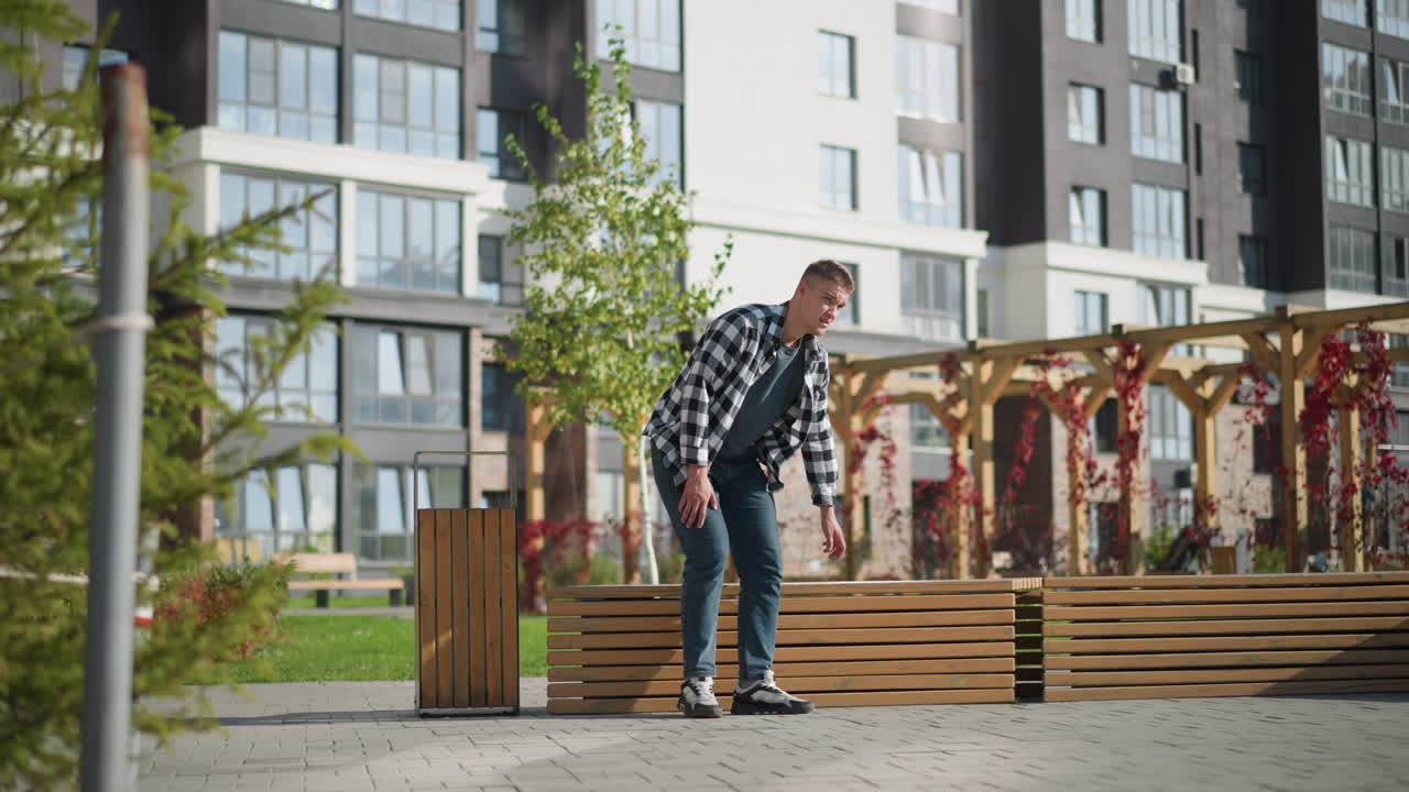 Young man reaches into front pocket for medicine while approaching wooden bench in sunny park. Foreground features pole and greenery, with decorated pergolas and tall buildings behind