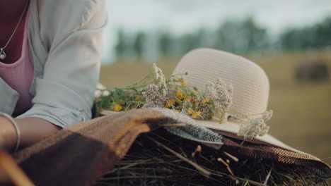 close-up of a sun hat and wildflowers resting on a blanket over a hay bale, with a soft-focus background of an open field and a partial view of a person