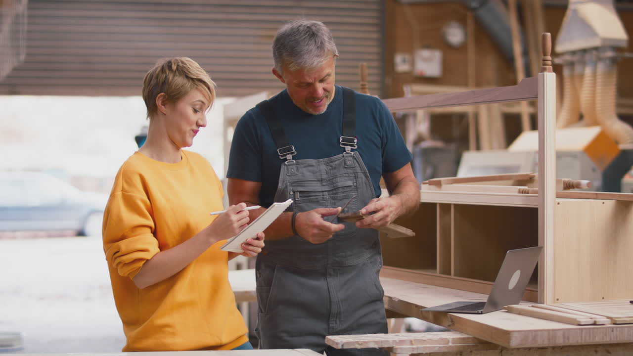 Female Apprentice Learning From Mature Male Carpenter With Laptop In Furniture Workshop