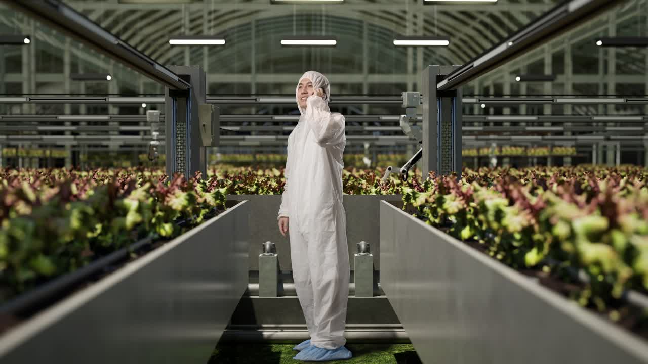 полный боковой вид тела азиатского исследователя, разговаривающего на смартфоне и улыбающегося, стоя в теплице с умными роботами-фермерами