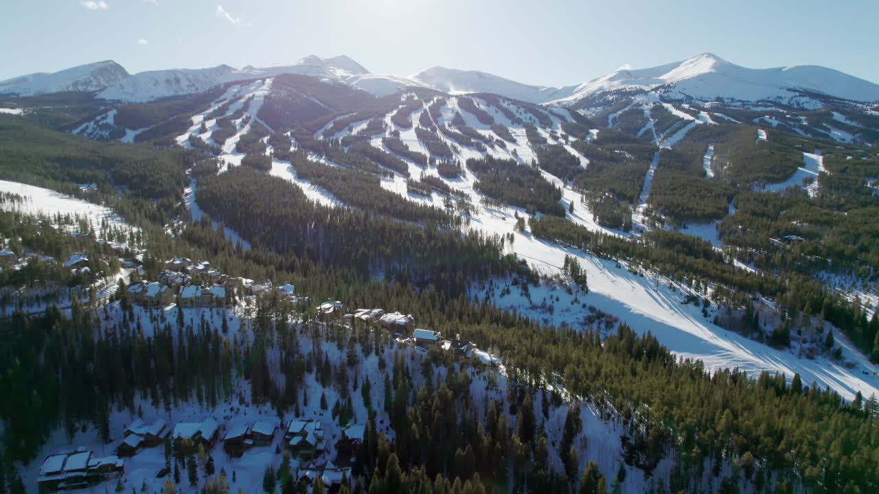 An epic aerial approaching the popular Breckenridge ski resort in Colorado, over a snow covered mountain ridge and high altitude trails full of winter skiers and snowboarders on a sunny winter day.