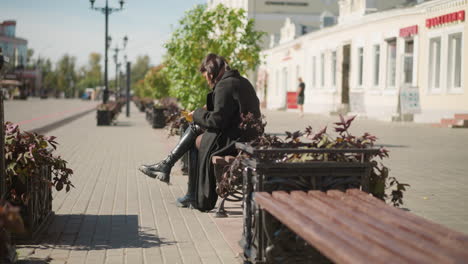 student sitting outdoors with crossed legs, reading a book and flipping through the pages, flowers sway in the wind, with blurred figures in the background during a sunny day