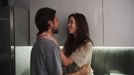 A-happy-brunette-girl-in-a-beige-T-shirt-hugs-her-brunette-boyfriend-with-stubble-in-a-blue-T-shirt-in-the-evening-in-a-modern-kitchen.-Happy-couple-guy-and-girl-hugging-during-their-romantic-evening-in-the-kitchen