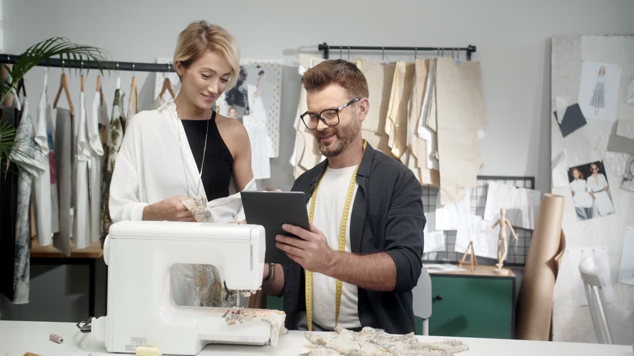 мужчина портной в очках сидит за столом со швейной машинкой, использует планшет и разговаривает с женщиной коллегой