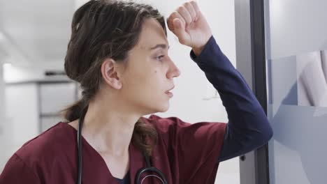 Doctora-Caucásica-Mirando-Por-La-Ventana-En-El-Pasillo-Del-Hospital,-Cámara-Lenta