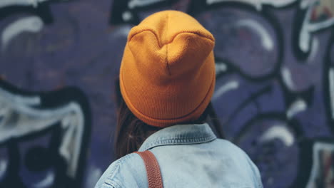 Back-View-Of-The-Close-Up-Of-The-Hipster-Stylish-Young-Girl-In-Hat-And-Sunglasses-Turning-Her-Head-Smiling-Cheerfully-To-The-Camera-On-The-Graffity-Background