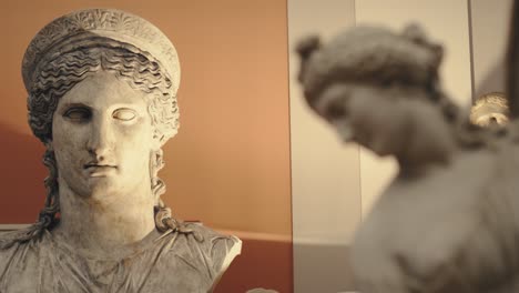 Ancient-antique-statue-of-a-young-woman-in-a-museum-with-moody-warm-lights-and-a-head-of-a-bigger-woman-statue-in-the-background