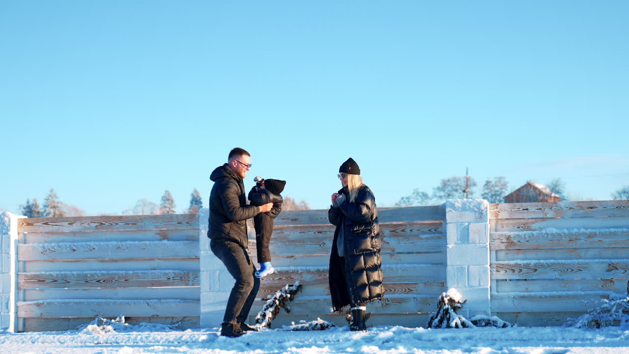 Cheerful father tosses his little toddler boy up. Mother stands beside clapping hands. Family outdoors in winter.
