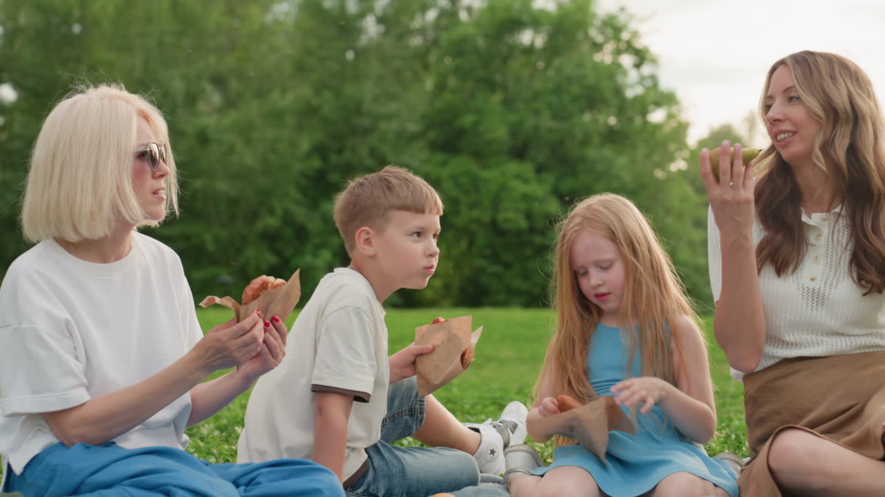 mothers and kids sitting on picnic mat in park enjoying snacks together, green grass background, casual summer outing, laughing and sharing food during family bonding time