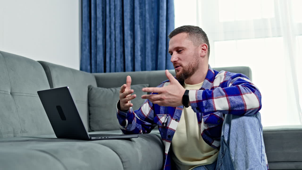 Un giovane uomo è seduto per terra e parla con qualcuno tramite un laptop in un soggiorno moderno durante il giorno.