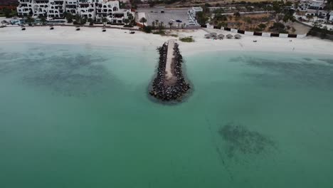 Un-Afloramiento-Rocoso-En-Aguas-Turquesas-En-La-Playa,-Playa-Caymancito,-Cerca-De-La-Paz,-En-Baja-California-Sur-Con-Complejos-Turísticos-Al-Fondo,-Vista-Aérea