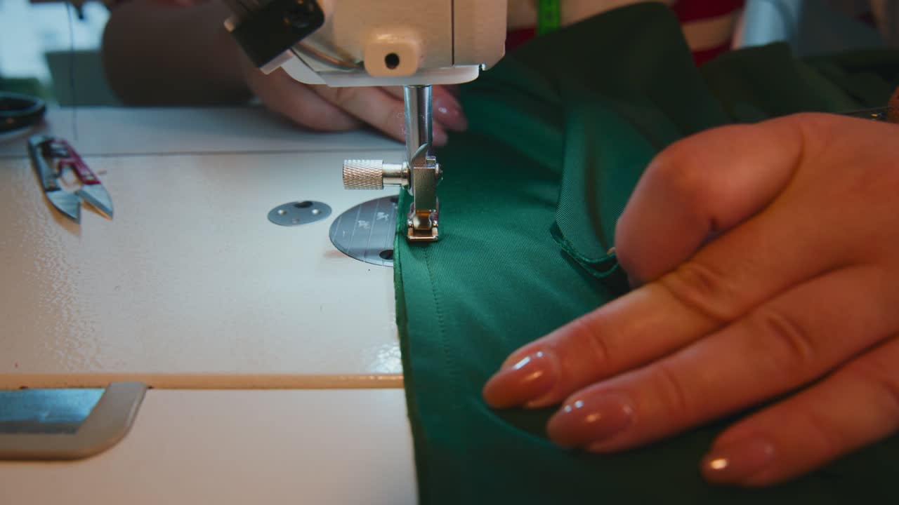 Primo piano di mani che guidano un tessuto verde sotto l'ago di una macchina da cucire. Le dita controllano delicatamente il materiale durante il processo di cucitura. L'attenzione rimane sul movimento preciso del cucito. Ideale per moda, tessile