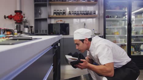 Mixed-race-male-chef-wearing-chefs-whites-in-a-restaurant-kitchen,taking-food-out-of-an-oven
