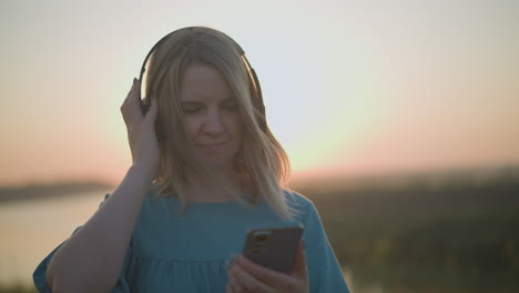 woman in a blue gown enjoys a peaceful sunset, wearing a black headset and using a smartphone, gently adjusting her headset with one hand