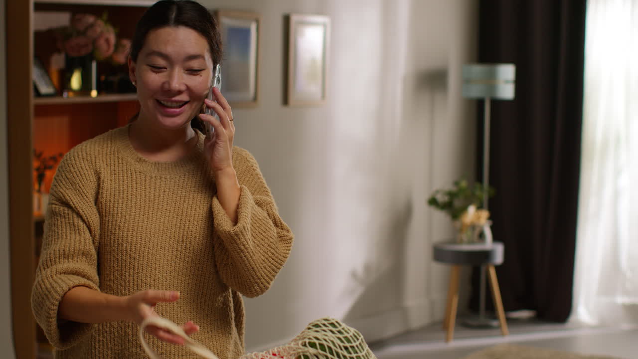 휴대 전화 로 이야기 하는 여자 가 음식 쇼핑 에서 집 으로 돌아오고 부 에 있는 카운터 에 신선 한 채소 가방 을 풀고 있다